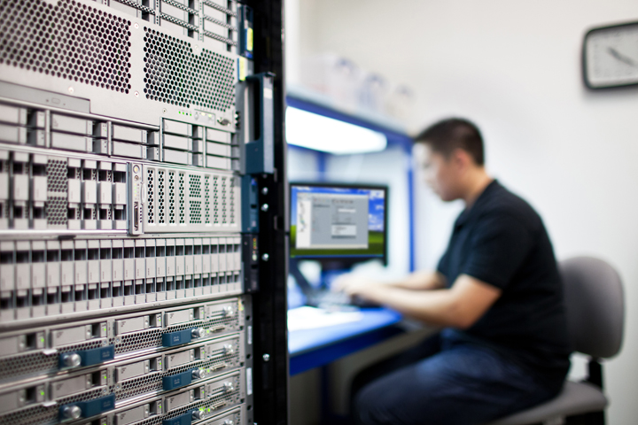 IT technician sits at a workstation beside a tall server rack, monitoring a monitor in a data center environment.