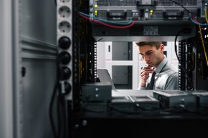 IT professional inspecting a server rack, looking at a laptop in a data center.