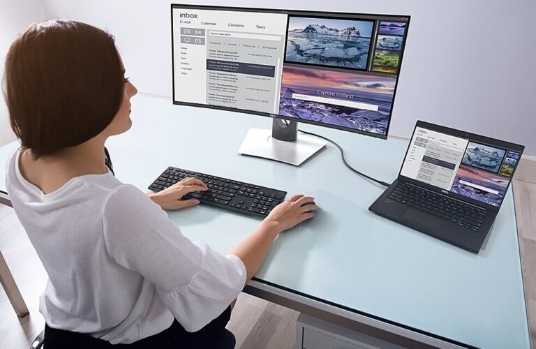 Person using a two-screen workstation: a large monitor and a connected laptop on a glass desk, displaying emails and photos side by side.