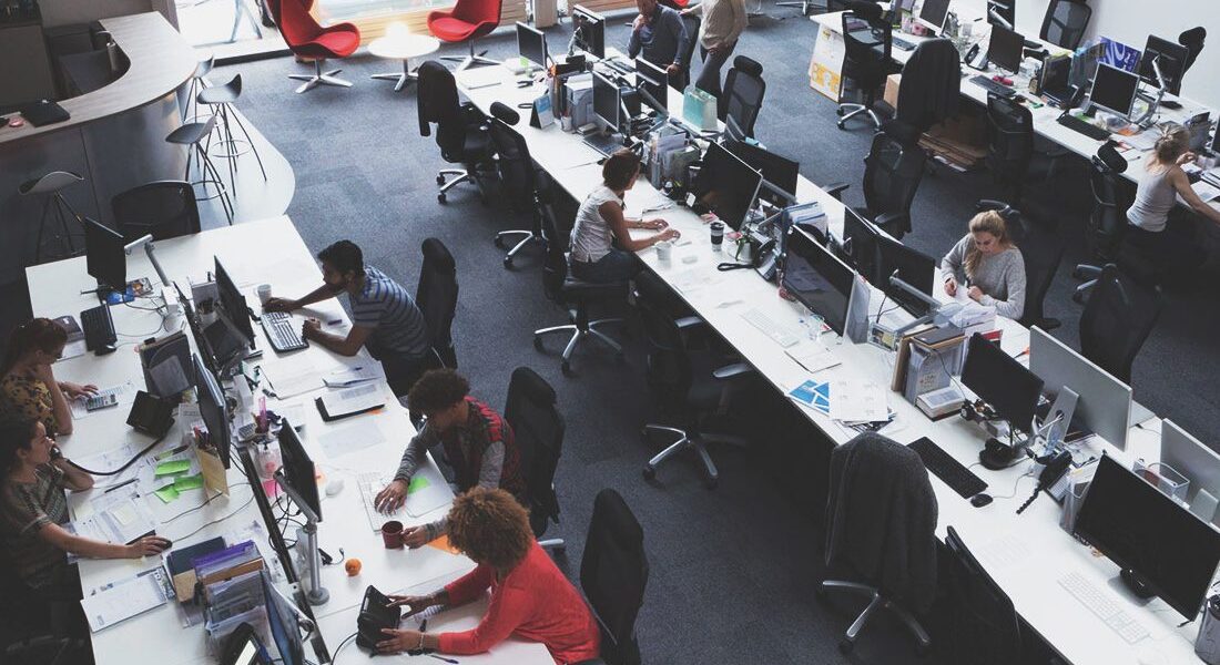 Open-plan office with long desks and multiple computer workstations; employees work at their computers amid large windows and scattered red lounge chairs in the background.