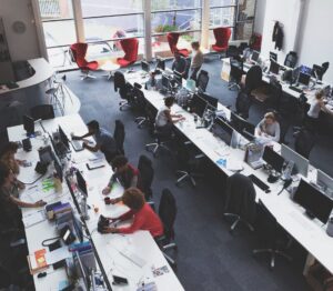 Open-plan office with long desks and multiple computer workstations; employees work at their computers amid large windows and scattered red lounge chairs in the background.