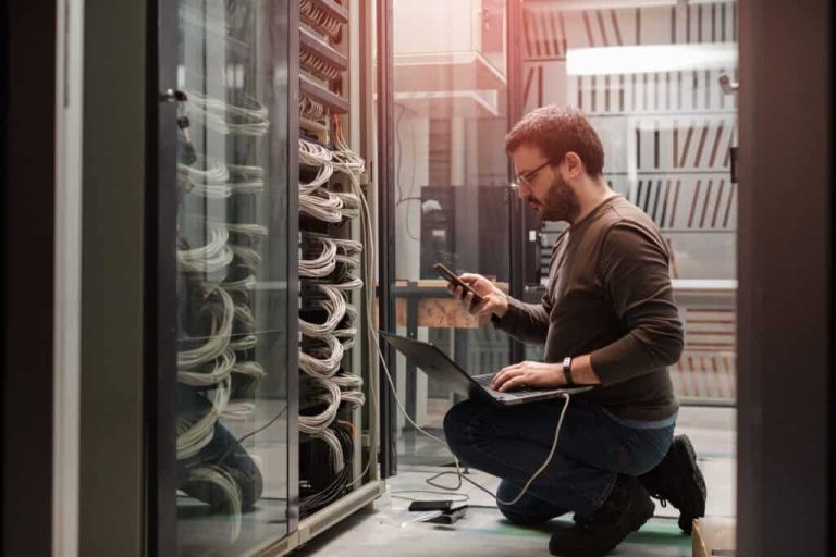 Technician kneeling beside server racks in a data center, using a laptop and smartphone.
